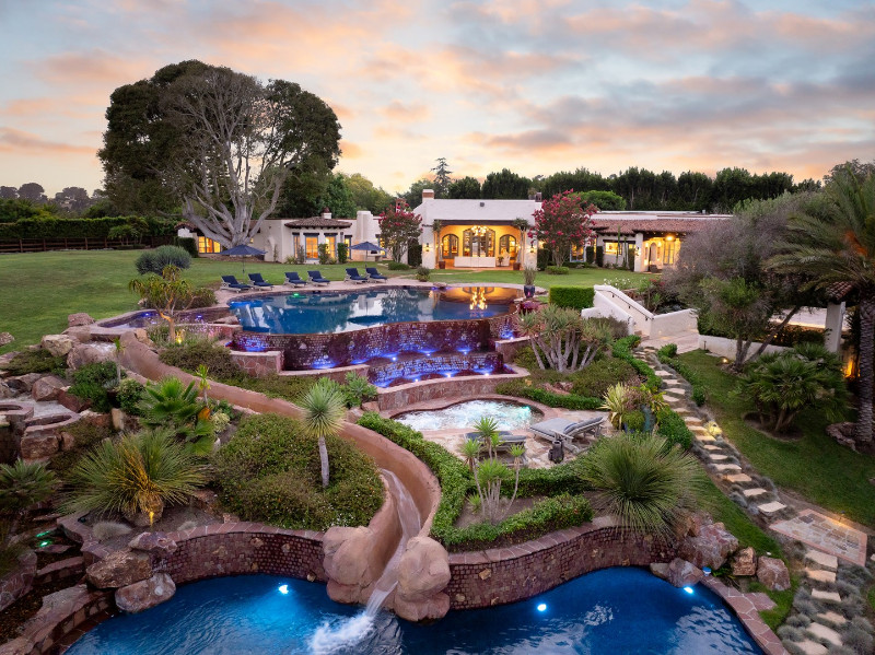 Luxury estate backyard with multi-tiered swimming pools featuring cascading waterfalls, a hot tub, and lush landscaping with tropical plants. The property includes a spacious lawn, elegant lounge chairs, and a Mediterranean-style villa with arched windows and warm lighting.